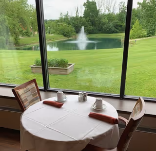 A round dining table covered with a white tablecloth set for two with cups, saucers, salt and pepper shakers, and folded red napkins. The table is positioned next to a large window overlooking a green lawn, a pond with a fountain, and trees in the background.