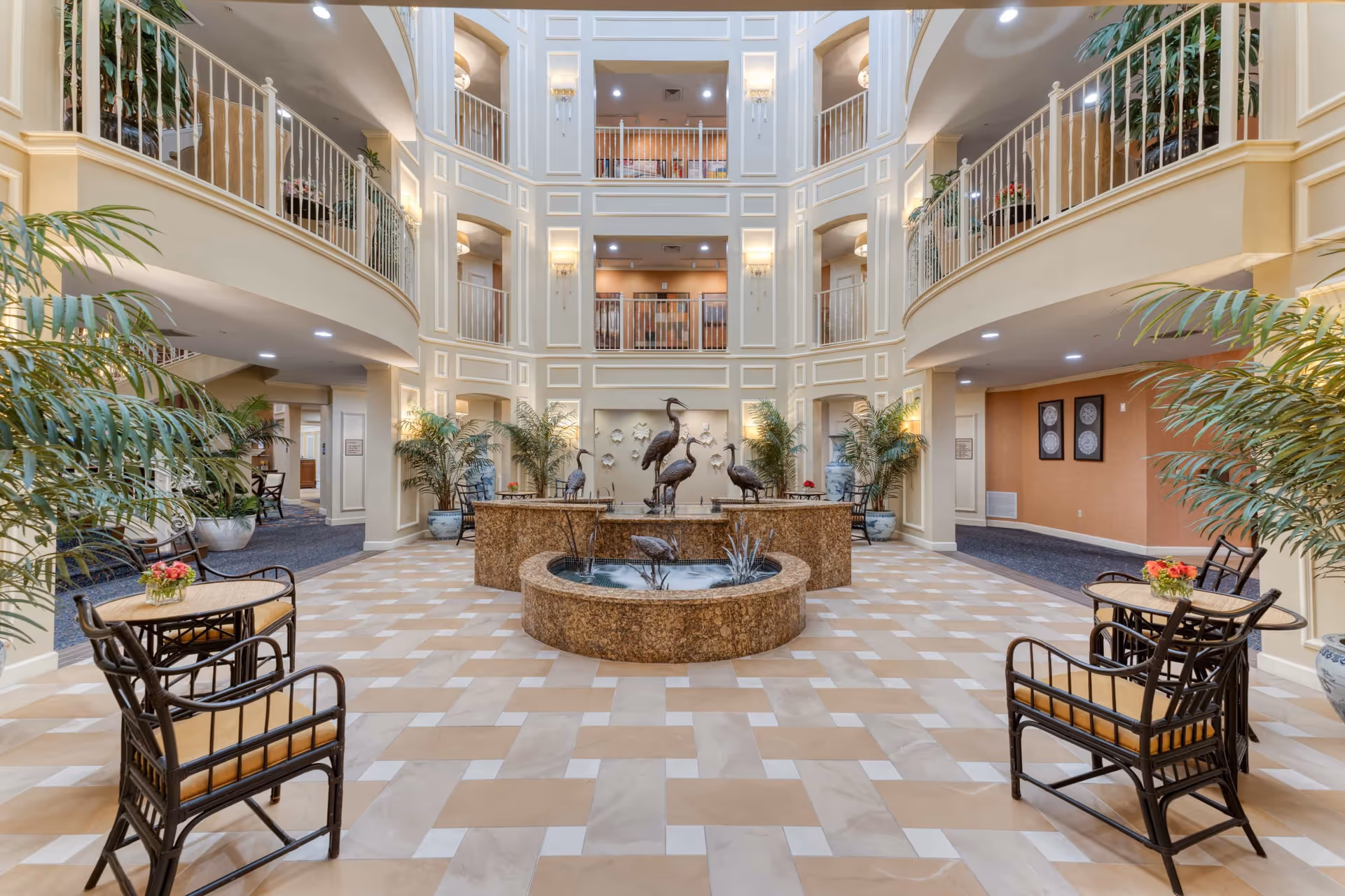 Spacious and elegant multi-story atrium with a central water fountain featuring bird sculptures, surrounded by potted plants and seating areas with tables and chairs. The area is well-lit with wall sconces and recessed lighting, and balconies overlook the open space from the upper floors.
