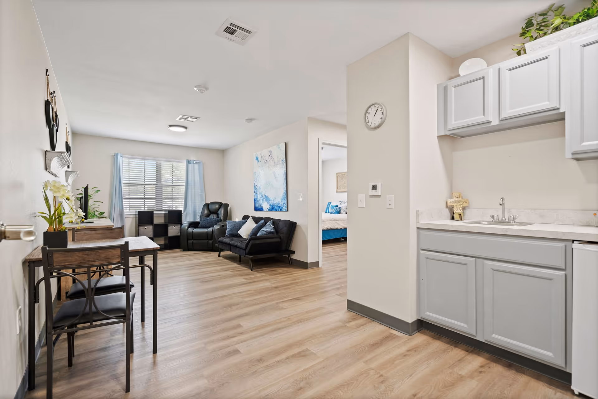 Interior view of a senior living apartment showing a small kitchenette with gray cabinets and a sink on the right, a living area with a black leather recliner and a black sofa with pillows, a small dining table with two chairs on the left, and a bedroom visible through an open doorway. The room has light-colored walls, wood flooring, a window with blue curtains, and a wall clock above the kitchenette.