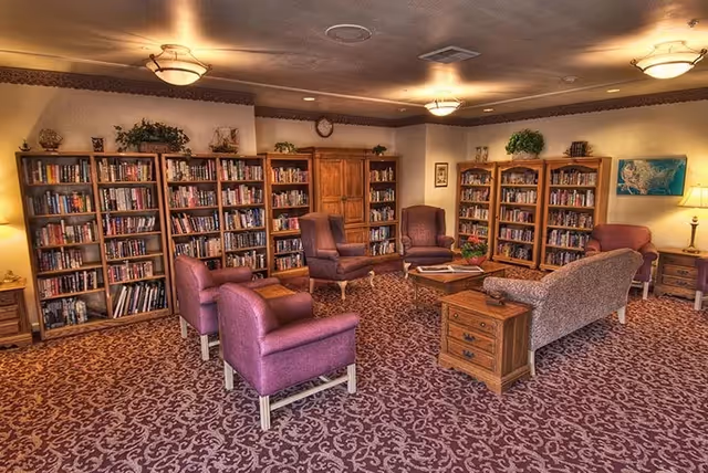 A cozy library or reading room with multiple bookshelves filled with books along the walls. The room features several comfortable upholstered chairs and a sofa arranged around a wooden coffee table. The carpet has a patterned design, and the room is softly lit by ceiling lights and a floor lamp. There are decorative plants on top of the bookshelves and framed pictures on the walls.