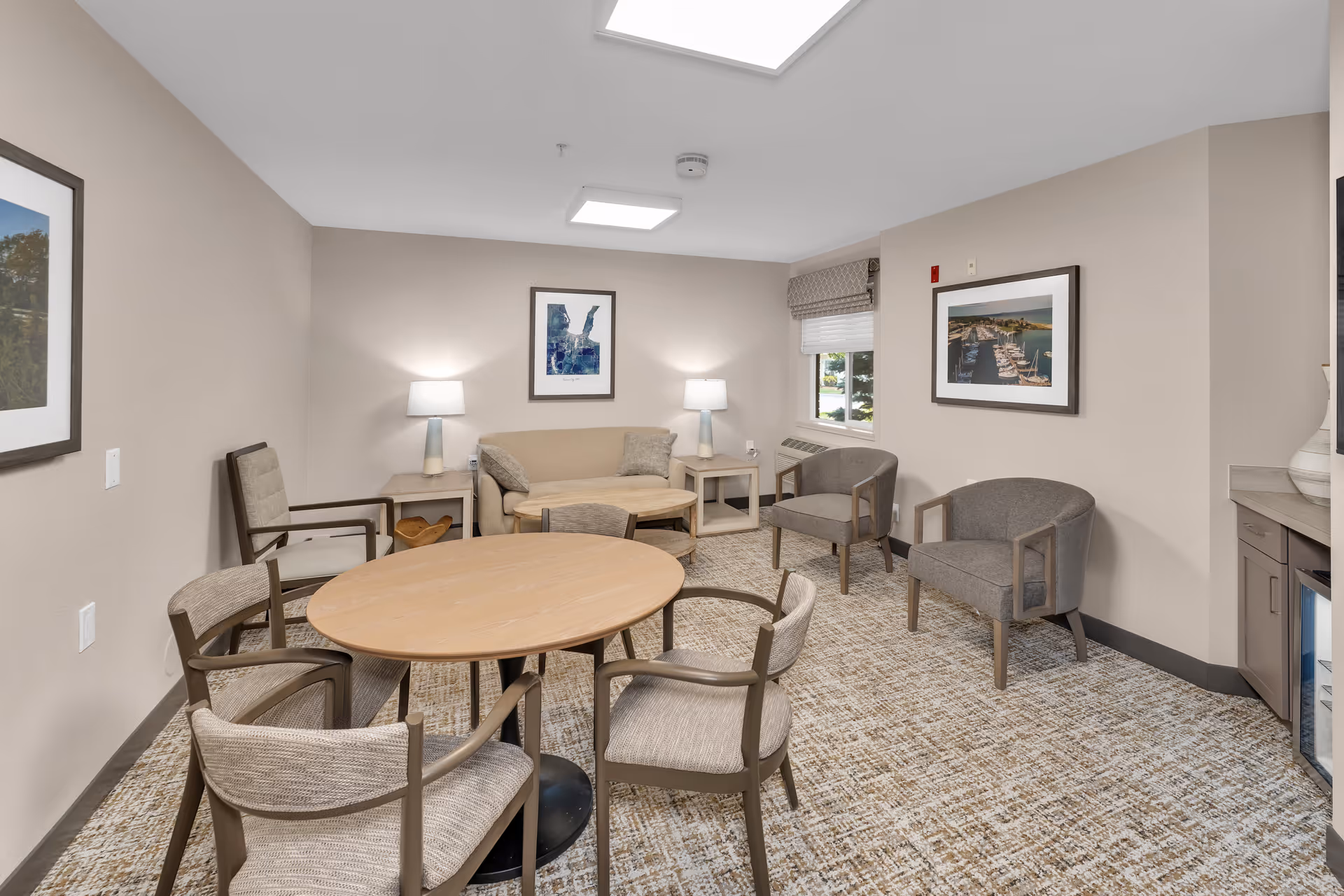 A cozy common area with a round wooden table surrounded by four chairs in the foreground. In the background, there is a beige sofa with two side tables each holding a lamp, two gray armchairs, and framed artwork on the walls. A window with a patterned valance lets in natural light. The room has neutral-colored walls and carpeted flooring.