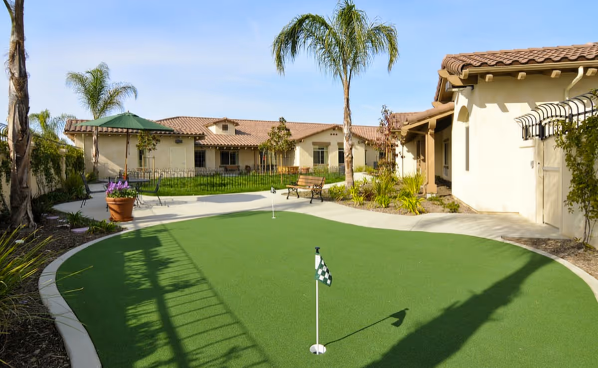 Outdoor courtyard area at ActivCare Bressi Ranch featuring a putting green with two golf holes and checkered flags, surrounded by a paved walkway, benches, potted plants, palm trees, and single-story buildings with tiled roofs under a clear sky.