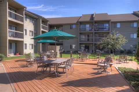 Outdoor patio area with wooden deck, several metal tables and chairs, and green umbrellas. The patio is surrounded by a multi-story residential building with balconies and windows. There is a small tree and some greenery near the patio.