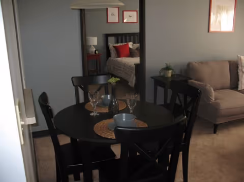 Interior view of a living space showing a round black dining table set with placemats, bowls, and glasses. Behind the table is a large mirror reflecting a bedroom with a bed, red pillows, and framed artwork. To the right of the dining area is a beige sofa with a pillow, and a small side table with a plant.