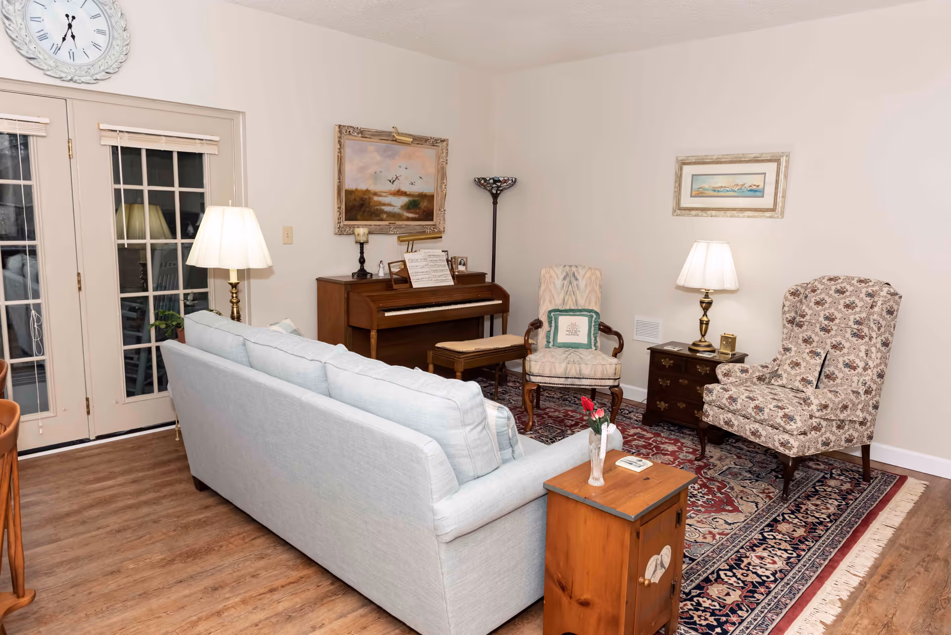 A cozy living room with a light blue sofa facing away, two patterned armchairs, a wooden piano with sheet music, a small wooden side table with a vase of flowers, two table lamps, a floor lamp, framed artwork on the walls, and French doors with blinds leading outside. The room has wooden flooring and a large patterned area rug.
