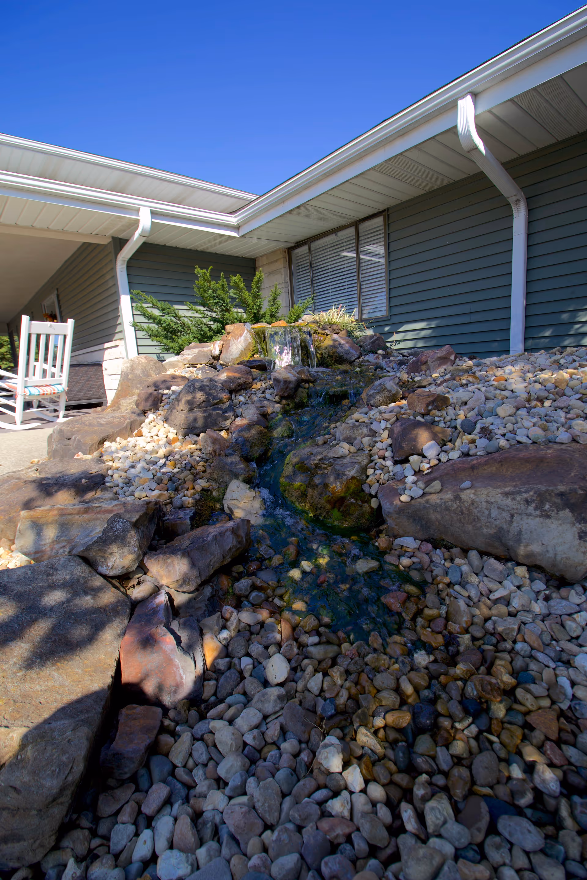 A rocky landscaped water feature and pebble bed in front of a single-story building with a rocking chair on the porch.