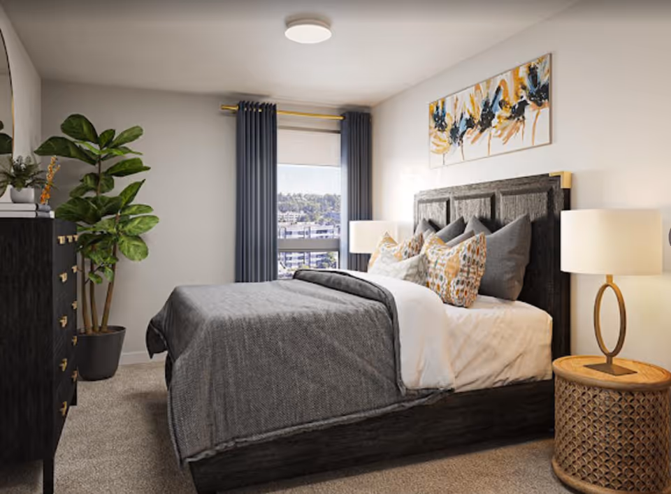 A cozy bedroom with a large bed featuring multiple pillows and a gray blanket. There is a wooden headboard, a round wooden nightstand with a modern lamp, a black dresser with gold handles, a large green potted plant, and a window with dark curtains letting in natural light. A colorful abstract painting hangs above the bed.