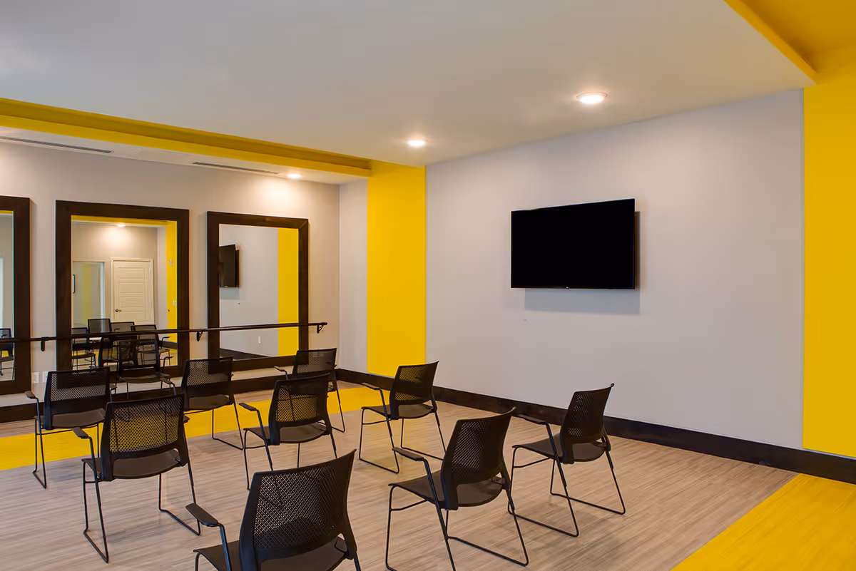 A modern room with yellow and gray walls featuring a row of black mesh chairs arranged facing a wall-mounted flat screen TV. The opposite wall has two large mirrors with dark wooden frames and a ballet barre beneath them. The floor is a combination of light wood and yellow accents.