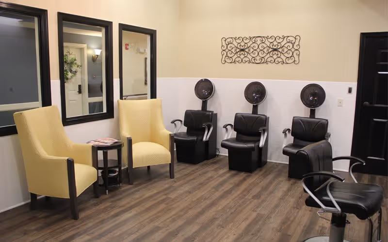 Interior salon area with two yellow armchairs, several black salon chairs and hooded hair dryers against a beige wall.