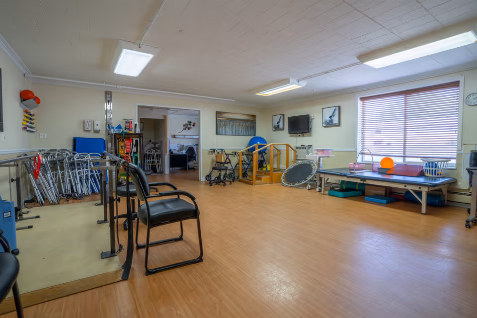 A spacious physical therapy room with wooden flooring and beige walls. The room contains various rehabilitation equipment including parallel bars, walkers, a small set of stairs with handrails, a trampoline, therapy balls, and a treatment table near a window with blinds. There are chairs and a wall-mounted TV, along with framed pictures on the walls.