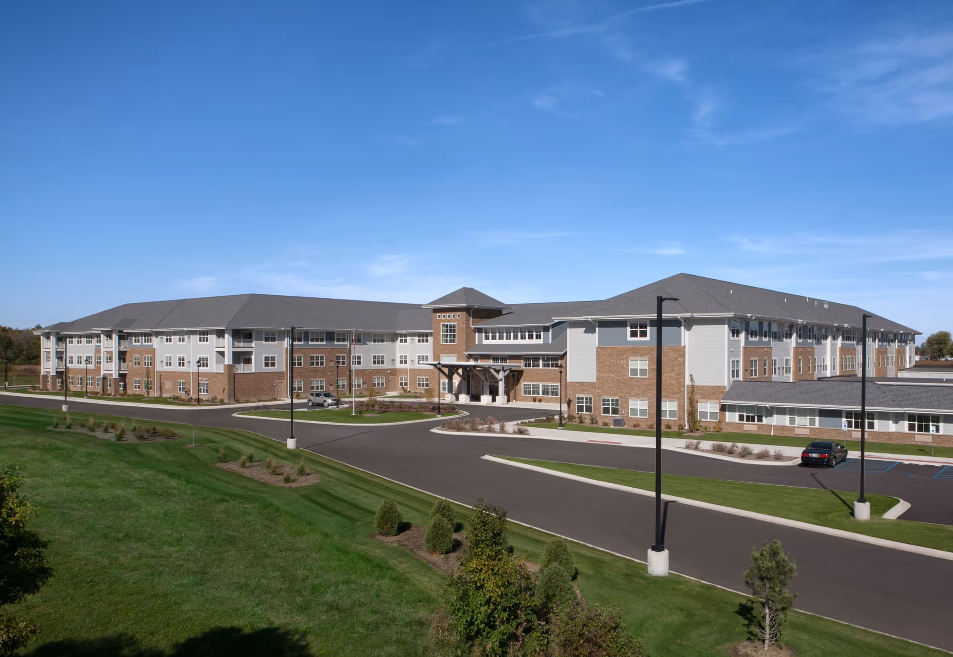 Exterior view of a large, modern senior living facility building with a combination of brick and light-colored siding, multiple windows, and a covered entrance. The building is surrounded by a paved driveway, landscaped green lawns, small trees, and street lamps under a clear blue sky.