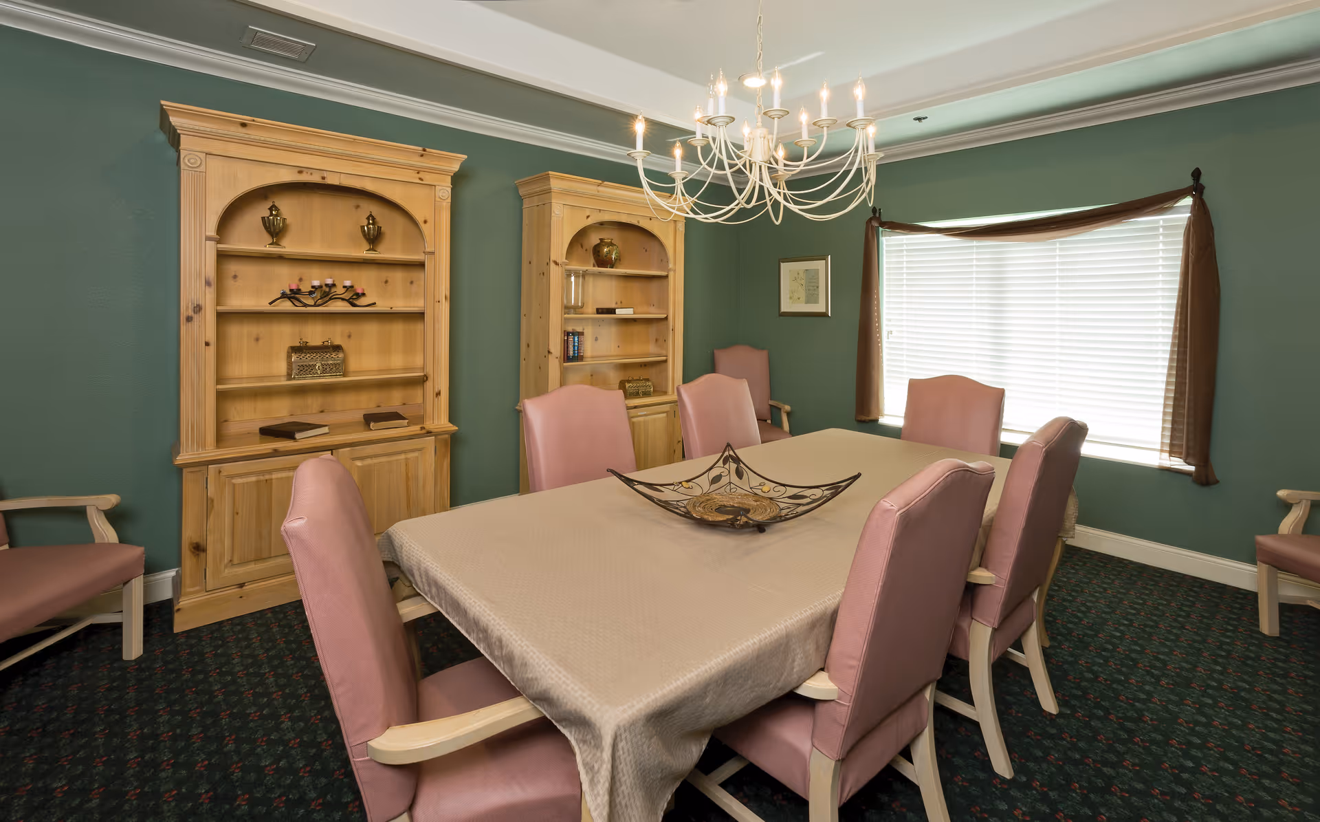A dining room with a rectangular table covered with a beige tablecloth surrounded by eight pink upholstered chairs. Two wooden bookshelves with decorative items are against a green wall. A chandelier hangs above the table and a window with closed blinds and brown curtains is on the right side.