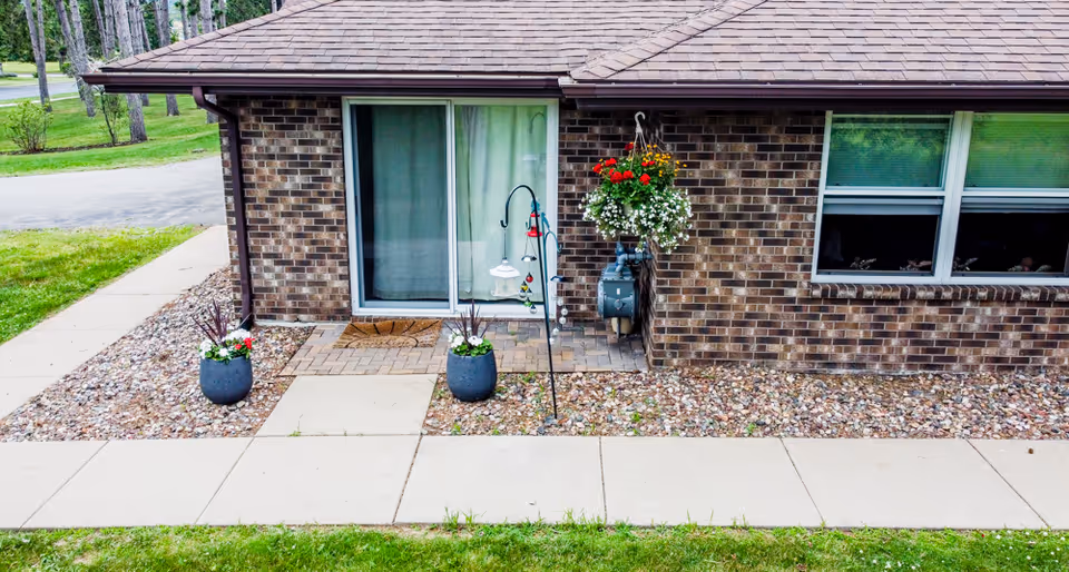 Exterior view of a brick building with a sliding glass door and a window. There are two large flower pots with plants on either side of the door, a hanging flower basket with red and white flowers, and a small paved area in front of the door. The building is surrounded by a sidewalk and grass.