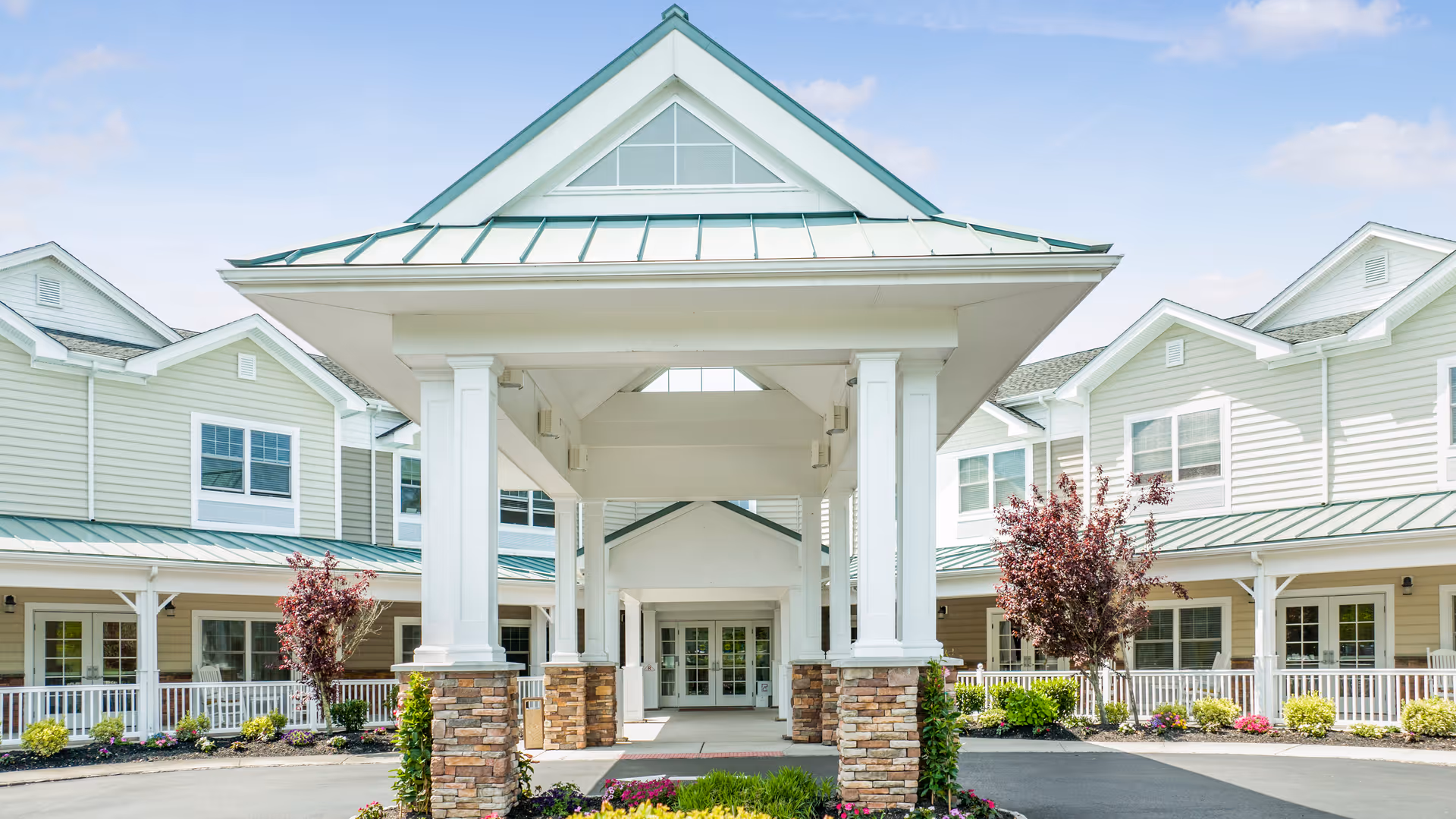 Front exterior view of The Terraces at Seacrest Village senior living facility with a covered entrance supported by white columns and stone bases, surrounded by well-maintained landscaping and a clear blue sky.