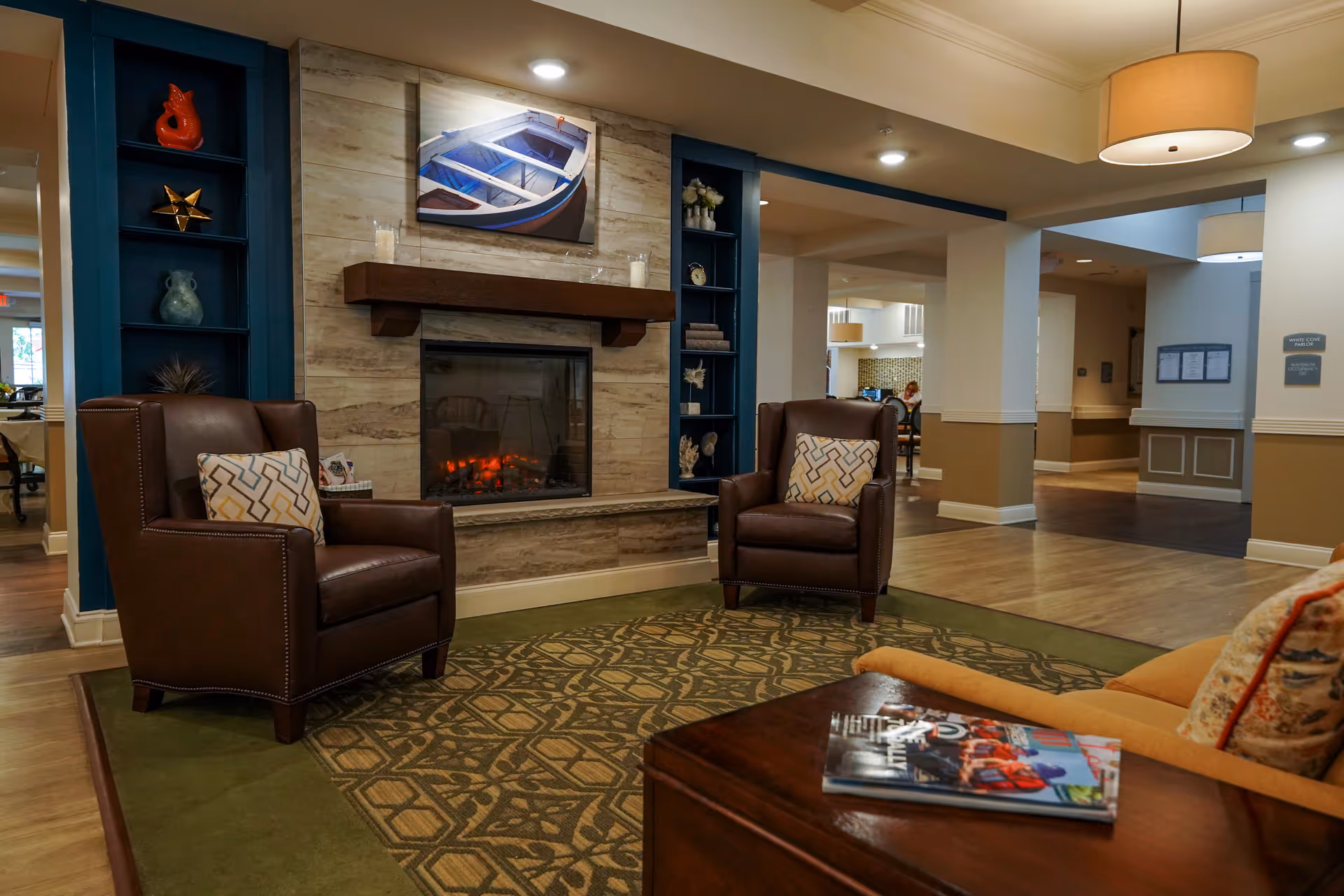 A cozy seating area in a senior living facility featuring two brown leather armchairs with patterned cushions, a fireplace with a wooden mantel, and a painting of a boat above it. The room has built-in shelves with decorative items, a green patterned rug, and a wooden coffee table with magazines. The background shows an open hallway and part of a dining area with a person seated.