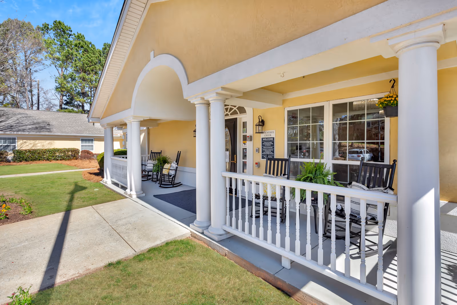 Front porch area of Peachtree Creek Memory Care assisted living facility in Georgia, featuring white columns, black rocking chairs with cushions, potted plants, and a yellow exterior wall with windows and a door under a covered entrance.