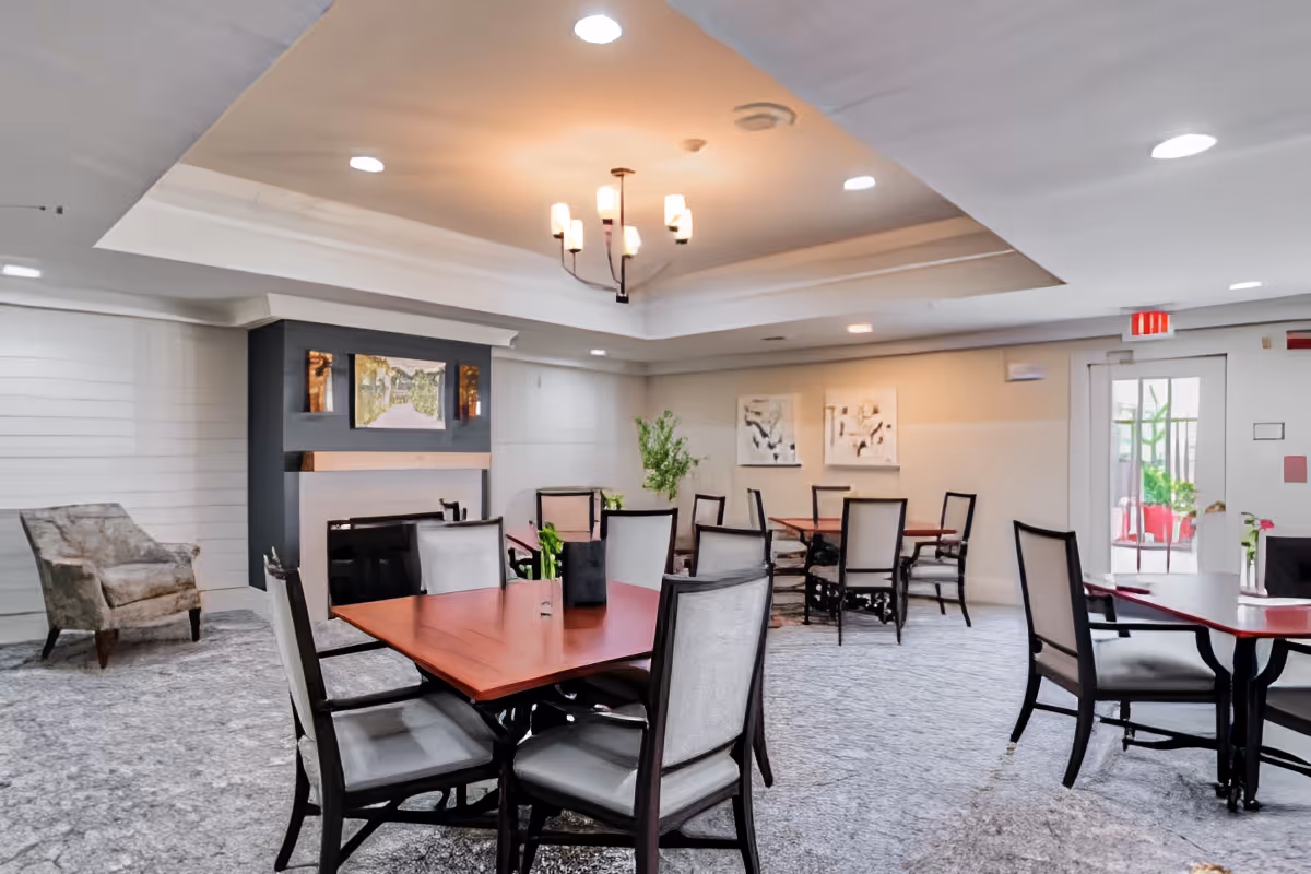 Well-lit communal dining area with multiple tables and chairs, a fireplace mantel, and overhead chandelier lighting.