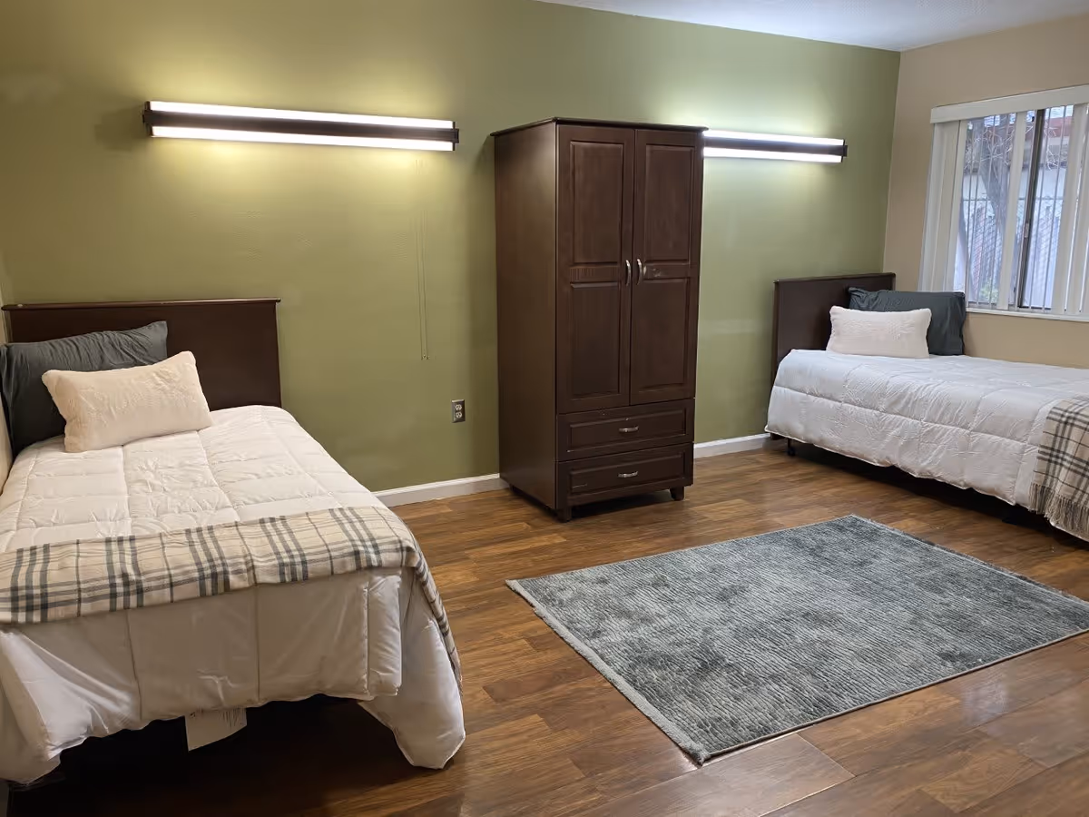 A simple bedroom with two single beds placed against opposite walls, each bed made with white bedding, a gray pillow, and a white decorative pillow. A dark wooden wardrobe is positioned between the beds against a green accent wall. The room has wooden flooring and a gray area rug in the center. Two modern horizontal light fixtures are mounted on the wall above each bed. A window with vertical blinds is visible on the right side.