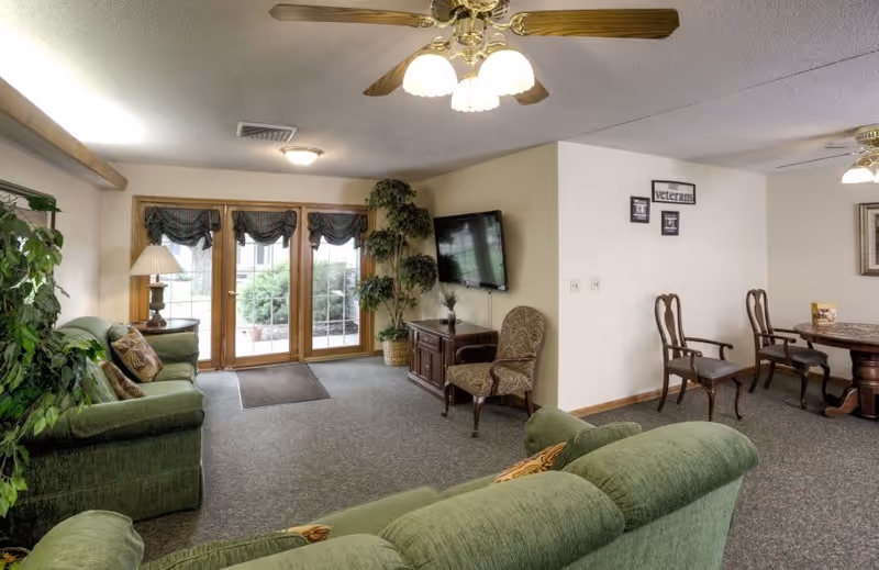 A cozy living room area with green upholstered sofas, a wooden TV stand with a flat-screen TV mounted on the wall, a patterned armchair, and a dining table with chairs. There are large glass doors with curtains leading outside, ceiling fans with lights, and several potted plants adding greenery to the space.