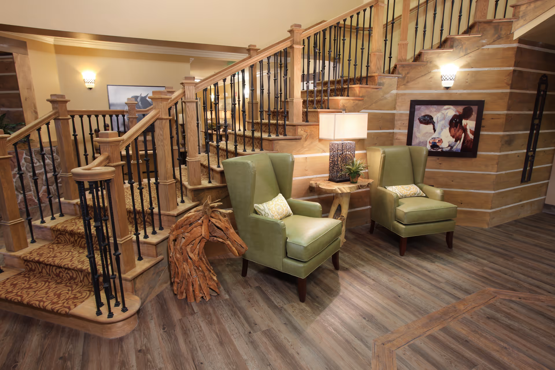 A cozy interior space featuring a wooden staircase with carpeted steps and black iron balusters. Two green upholstered armchairs with decorative pillows are positioned next to a small wooden side table with a lamp and a small plant. The walls are wooden with horizontal paneling, and there is artwork on the wall including a painting of a cow. The floor is wood with a natural finish.