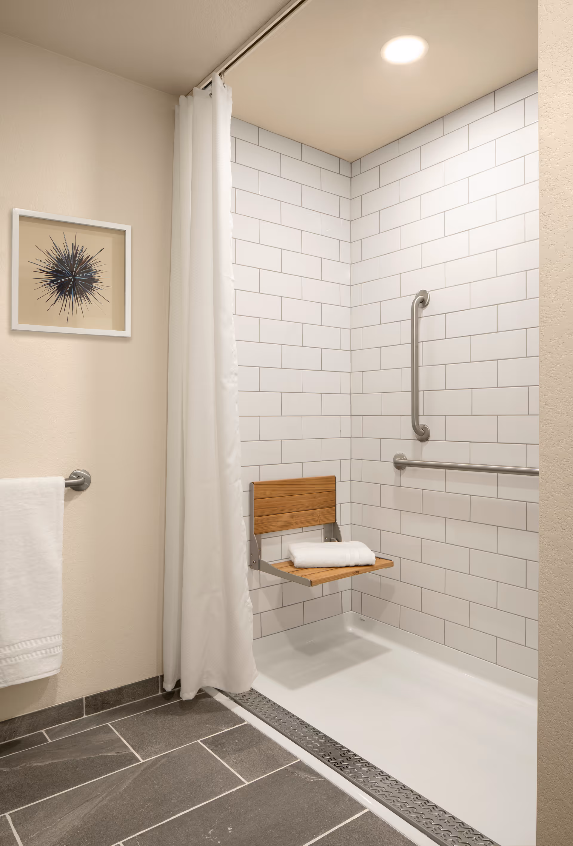 Accessible walk-in shower with white subway tile, stainless grab bars, a fold-down wooden seat holding folded towels, and a white shower curtain.
