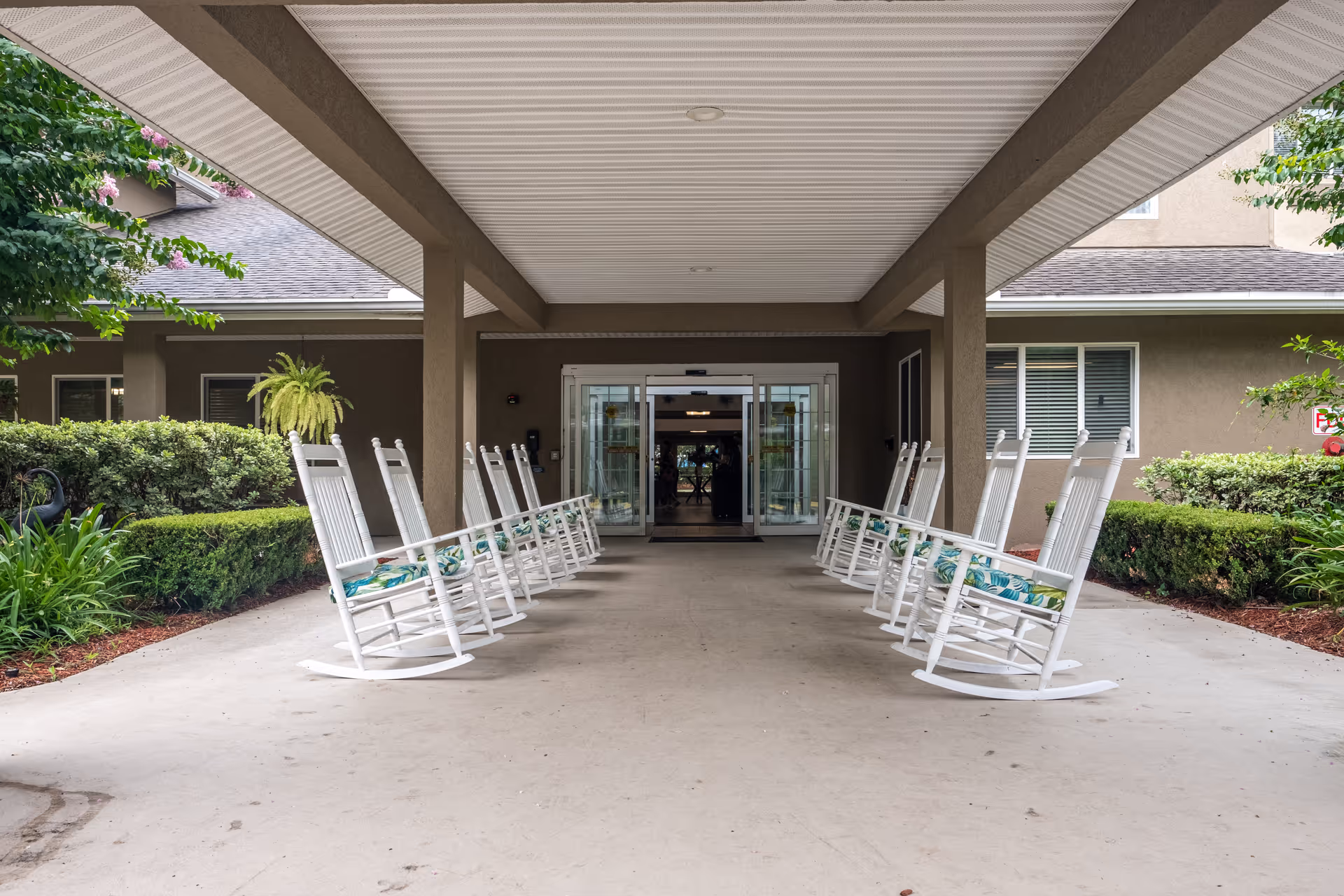 Covered entrance area of a senior living facility with two rows of white rocking chairs facing each other. The entrance has automatic sliding glass doors and is surrounded by neatly trimmed bushes and greenery.
