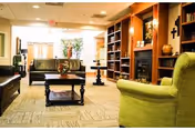 A cozy living room area with green upholstered chairs and a dark wooden coffee table. There are built-in wooden bookshelves with decorative items and a fireplace. The room is well-lit with natural light coming from the windows in the background.