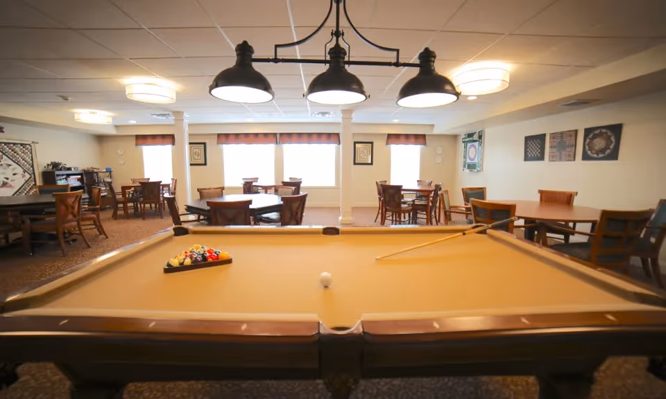 A recreational room with a pool table in the foreground, set up with billiard balls and a cue stick. In the background, there are multiple tables and chairs arranged for seating, with framed artwork on the walls and large windows letting in natural light. The ceiling has several light fixtures, including a three-light pendant above the pool table.