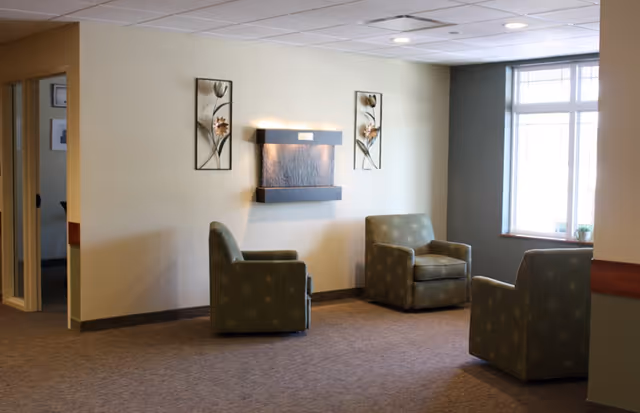 A quiet seating area in a memory care facility with three green upholstered armchairs arranged around a wall-mounted water feature. The walls are light-colored with two decorative floral metal wall hangings on either side of the water feature. A large window on the right lets in natural light, and the floor is carpeted.