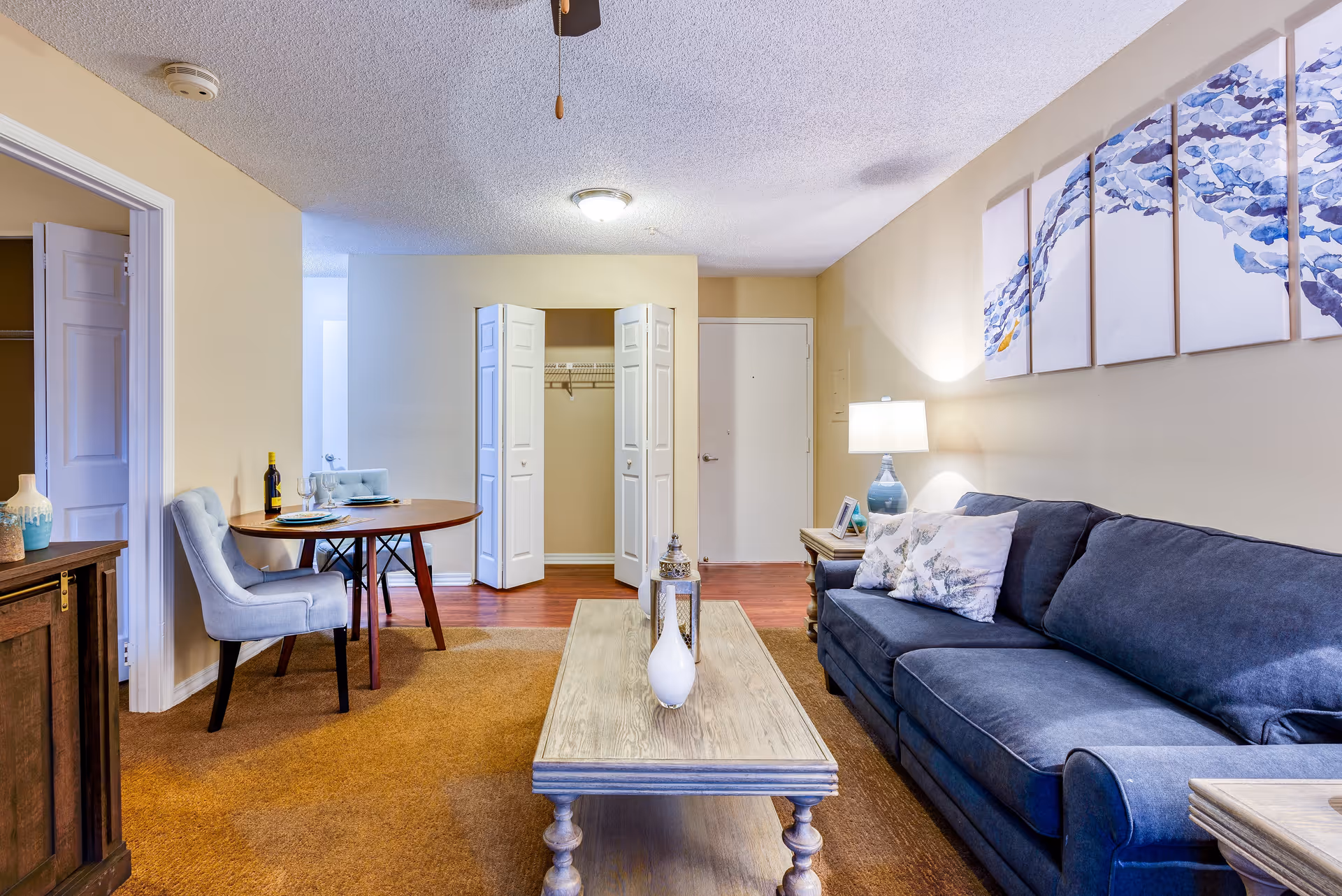 A cozy living room area in a senior living facility with a dark blue couch adorned with decorative pillows, a wooden coffee table with a white vase and lantern, a side table with a lamp, and a round dining table with two light blue upholstered chairs. The walls are light beige with a multi-panel blue abstract artwork above the couch. There is an open closet with white bi-fold doors and a white entrance door in the background.