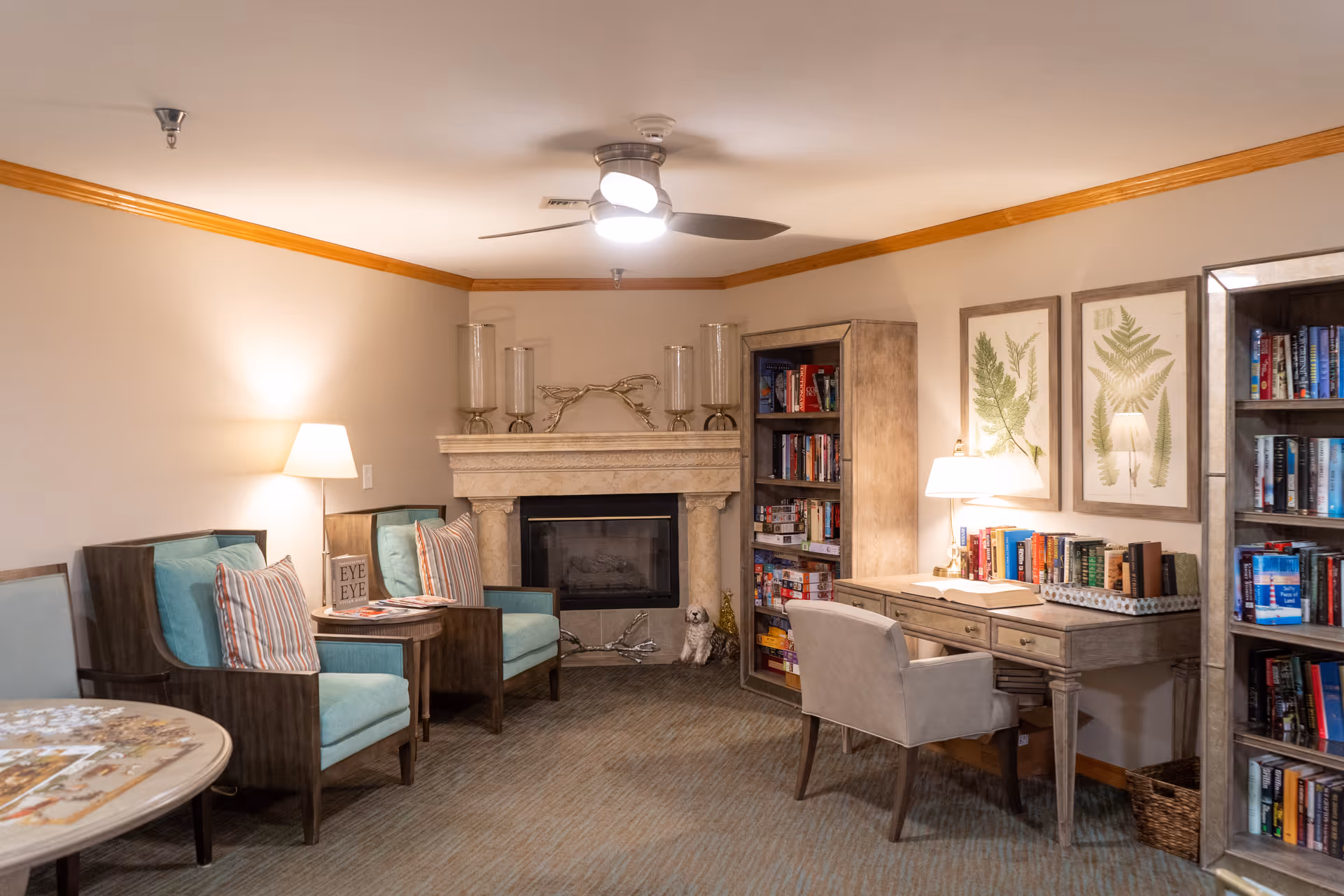 A cozy living room area in a senior living facility with two turquoise armchairs with striped pillows, a small round table with magazines, a stone fireplace with decorative candle holders, a bookshelf filled with books and board games, a desk with a lamp and books, two framed botanical prints on the wall, and a ceiling fan with a light.