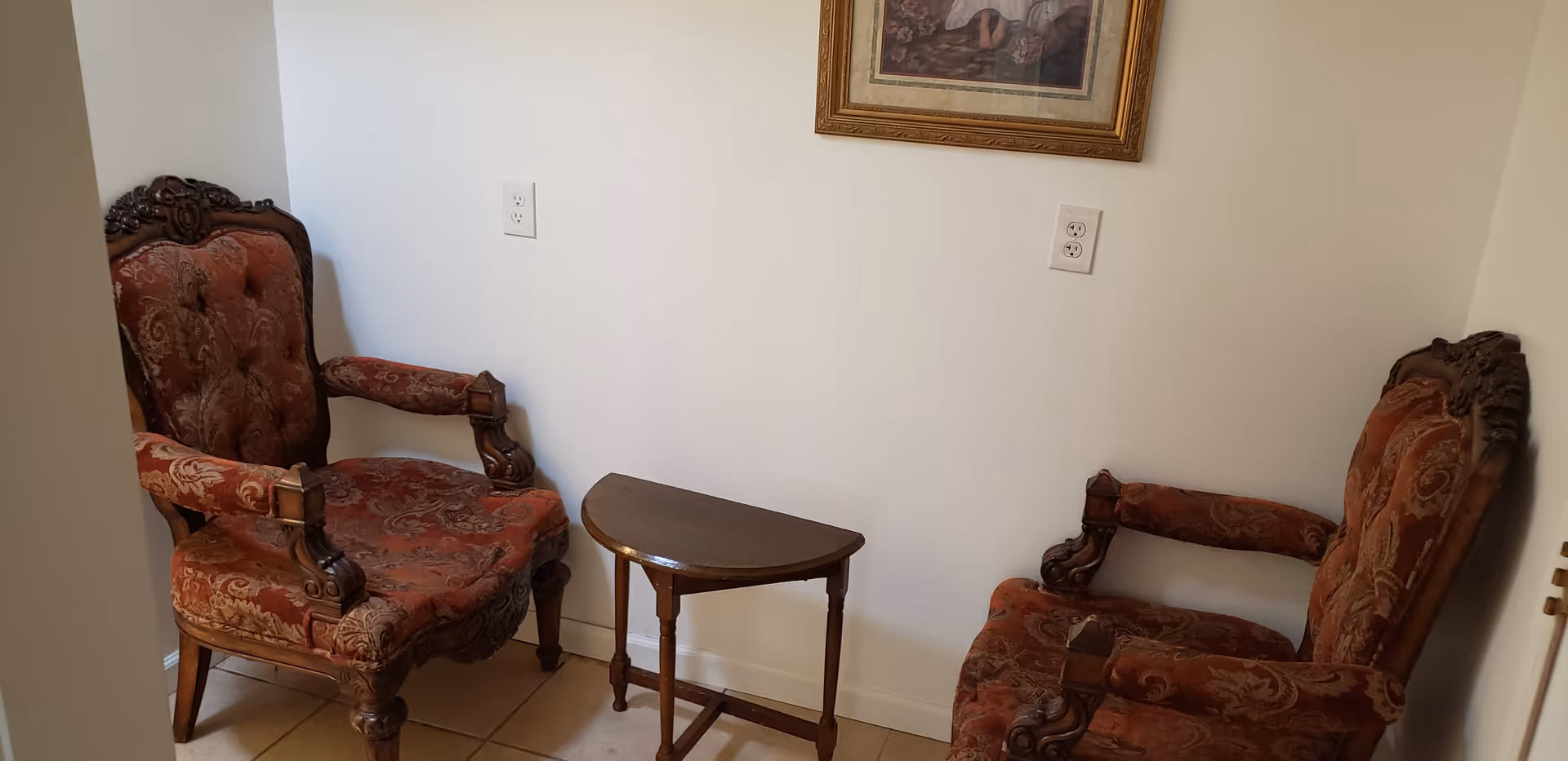Two ornate wooden armchairs with red patterned upholstery facing each other with a small wooden half-moon table between them, set against a plain white wall with two electrical outlets and a framed picture above.