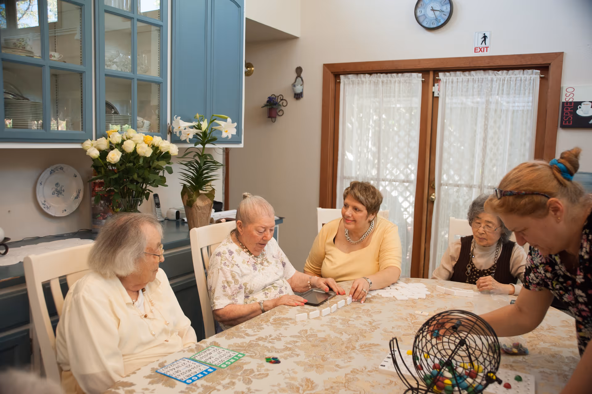 A group of elderly women and a caregiver sitting around a table playing a game with bingo cards and numbered tiles in a cozy room with blue cabinets, flowers, and a glass door with curtains.