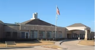 Exterior view of a single-story senior living facility building with a circular driveway, an American flag on a flagpole in front, and clear blue sky.