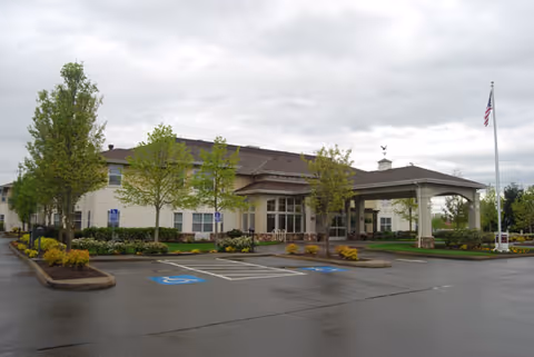 Front entrance of a single-story senior living building with a covered driveway, flagpole, landscaping, and parking spaces.