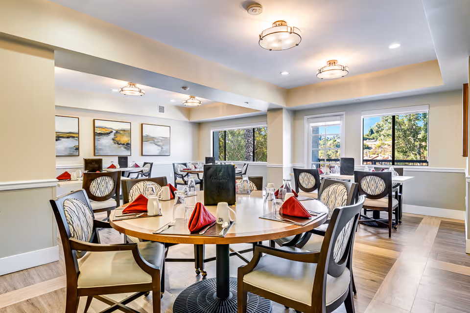 Bright and spacious dining room in a senior living facility with round and square tables set with red folded napkins, glasses, and menus. The room features large windows with views of greenery outside, light-colored walls, wooden flooring, and modern ceiling lights. Framed landscape paintings hang on the wall.