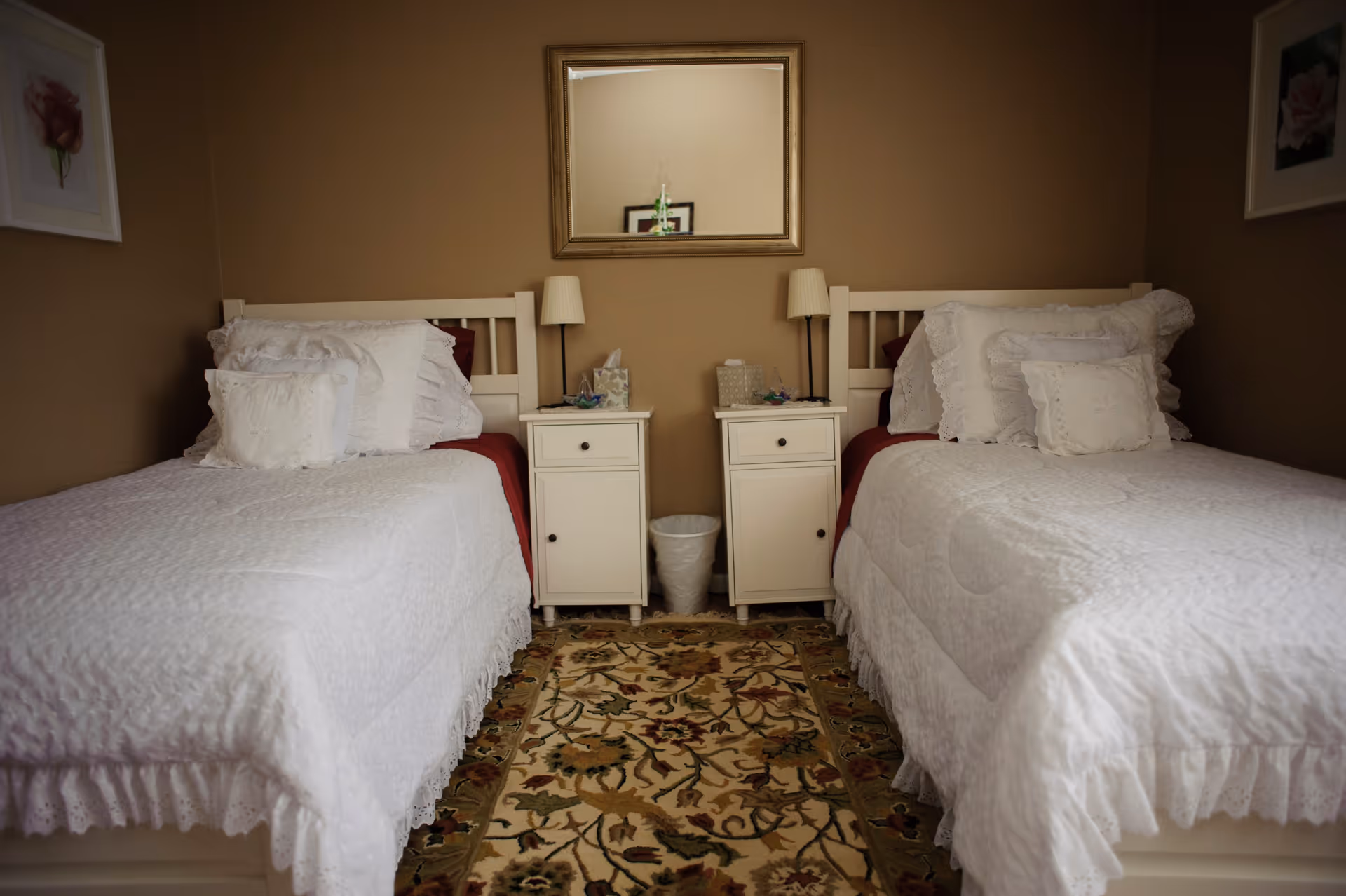 A cozy bedroom with two twin beds placed parallel to each other, each bed dressed in white bedding with decorative pillows. Between the beds are two white nightstands with lamps, a tissue box, and other small items. A floral patterned rug lies on the floor, and a rectangular mirror hangs on the beige wall above the nightstands. Framed floral artwork is visible on the side walls.