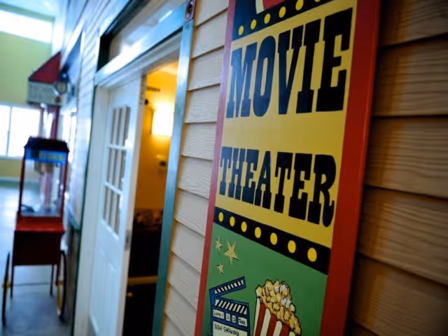 Interior hallway with a colorful sign on the wall that reads 'Movie Theater' featuring popcorn and film reel graphics, and a popcorn machine visible further down the hallway.