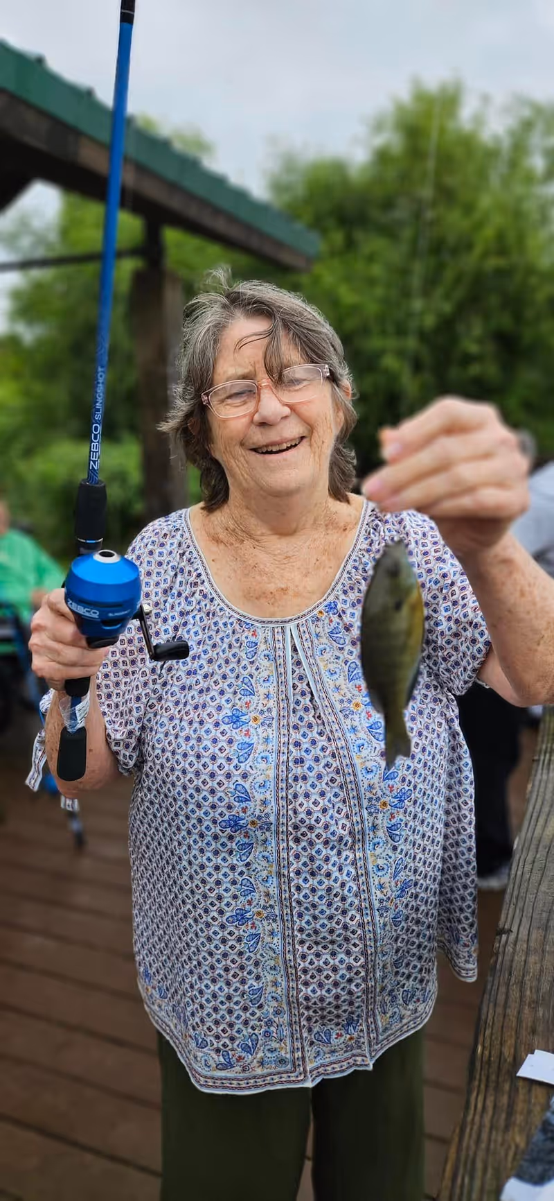 An elderly woman smiling and holding a fishing rod in one hand and a small fish in the other, standing on a wooden deck outdoors with greenery in the background.