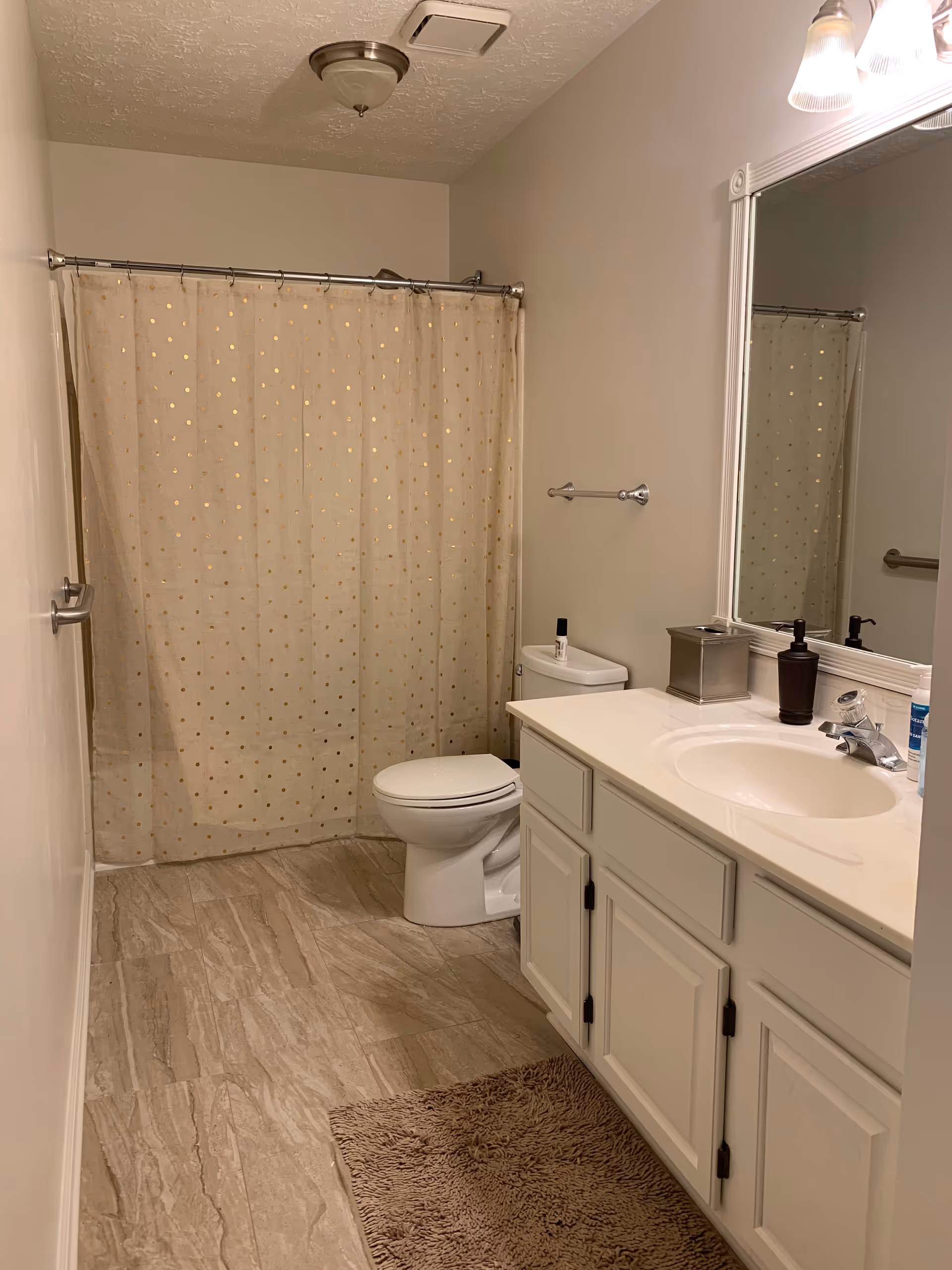 A clean bathroom with a white toilet, a white vanity with a sink, a large mirror above the sink, and a shower with a beige curtain featuring gold polka dots. The floor has beige tiles and there is a brown bath mat in front of the vanity.
