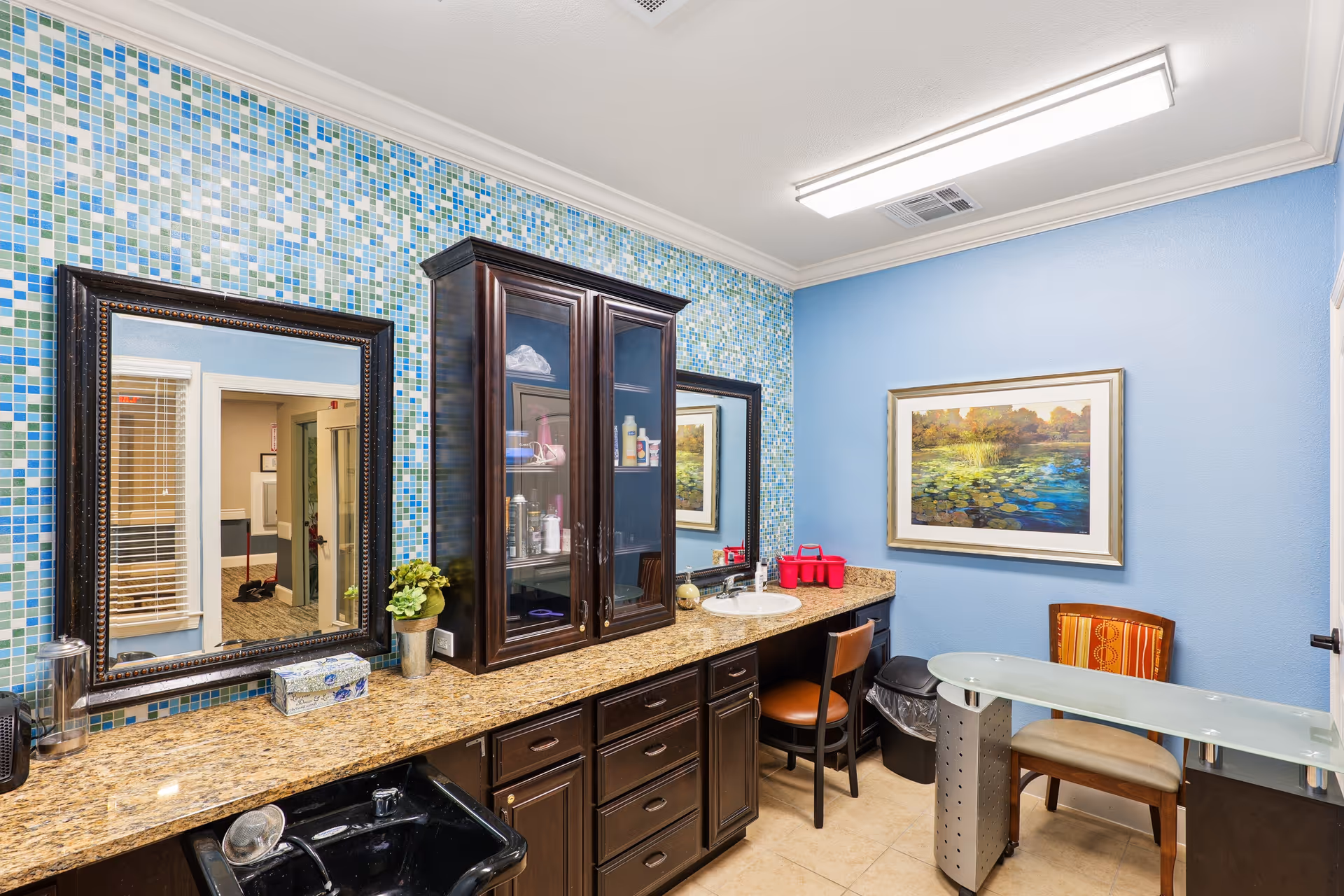 Bright interior grooming area with a mosaic tile wall, granite countertop with sinks, dark wood cabinets, mirrors and seating.