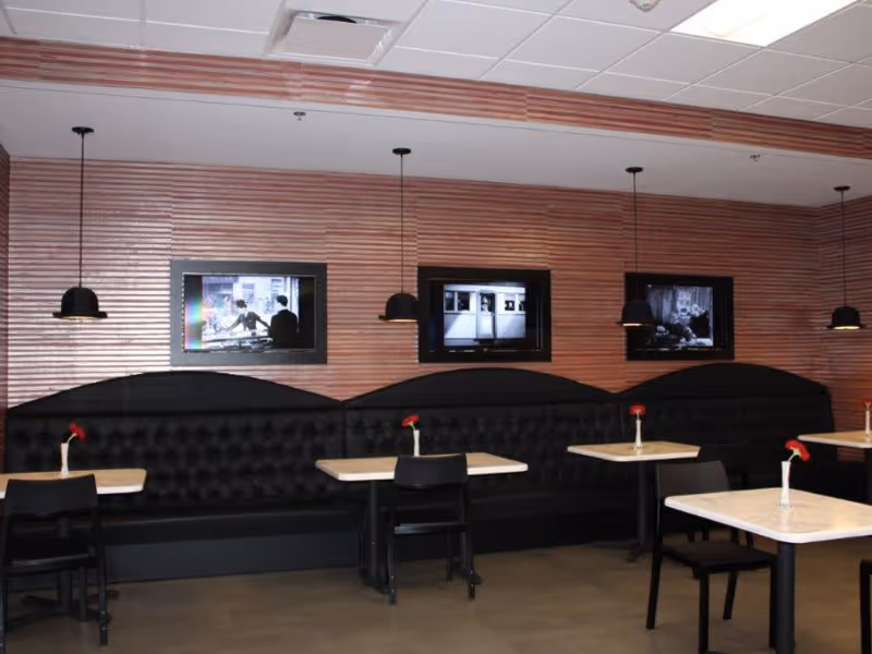 Interior of a dining area with black cushioned bench seating along a wooden paneled wall. There are three small square tables with black chairs, each table having a single red flower in a white vase. Above the bench seating, three framed black and white pictures are displayed on the wall. Four black pendant lights hang from the ceiling above the tables.