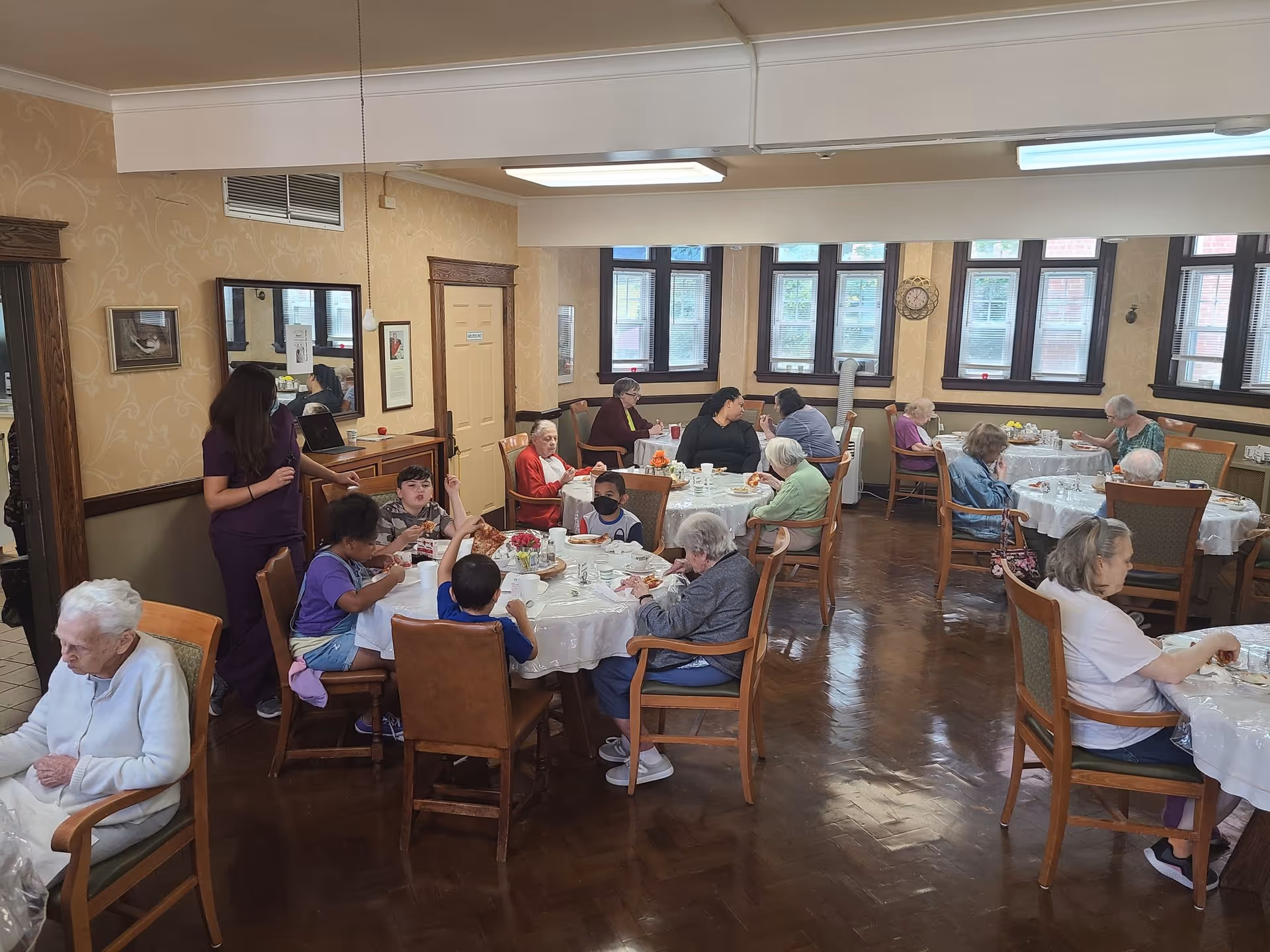 A dining room in a senior living facility with several elderly residents and a few children seated at round tables covered with white tablecloths, eating and socializing. A staff member in purple scrubs is standing near one of the tables. The room has wooden floors, beige patterned wallpaper, and multiple windows letting in natural light.