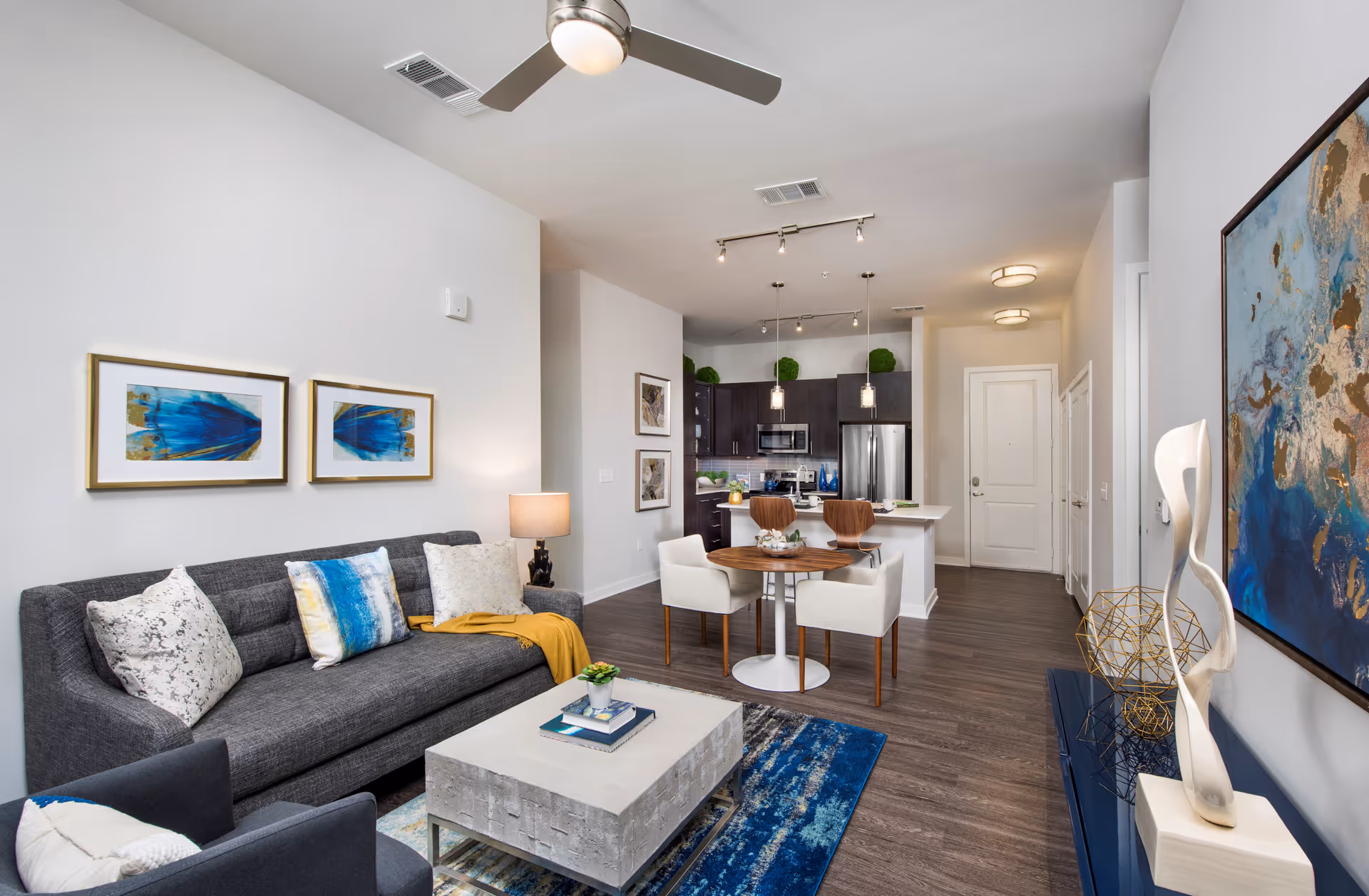 Modern living room and kitchen area with a gray sofa adorned with decorative pillows, a concrete coffee table on a blue patterned rug, a round wooden dining table with four upholstered chairs, and a kitchen with dark cabinets, stainless steel appliances, and pendant lighting. The room features white walls with framed artwork and a ceiling fan.