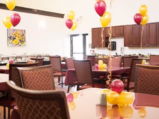 A decorated dining room with multiple tables and chairs. Each table has maroon and yellow balloons tied to small balloon weights. The room has wooden cabinets along one wall and a framed picture with sunflowers. There is a door with an exit sign above it in the background.