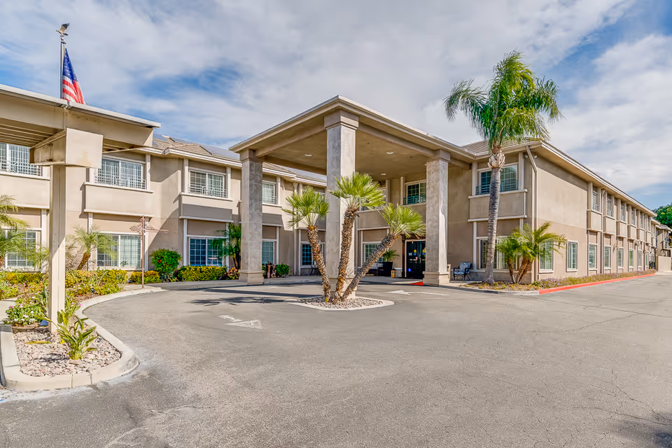 Two-story beige senior living facility with porte-cochère, palm trees, and circular driveway.