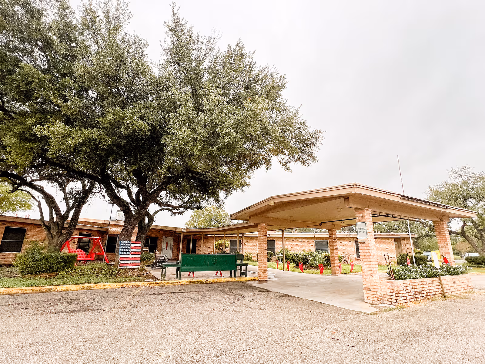 Front view of a single-story brick senior living facility with a covered drop-off/portico, large trees, benches and an American flag decoration.
