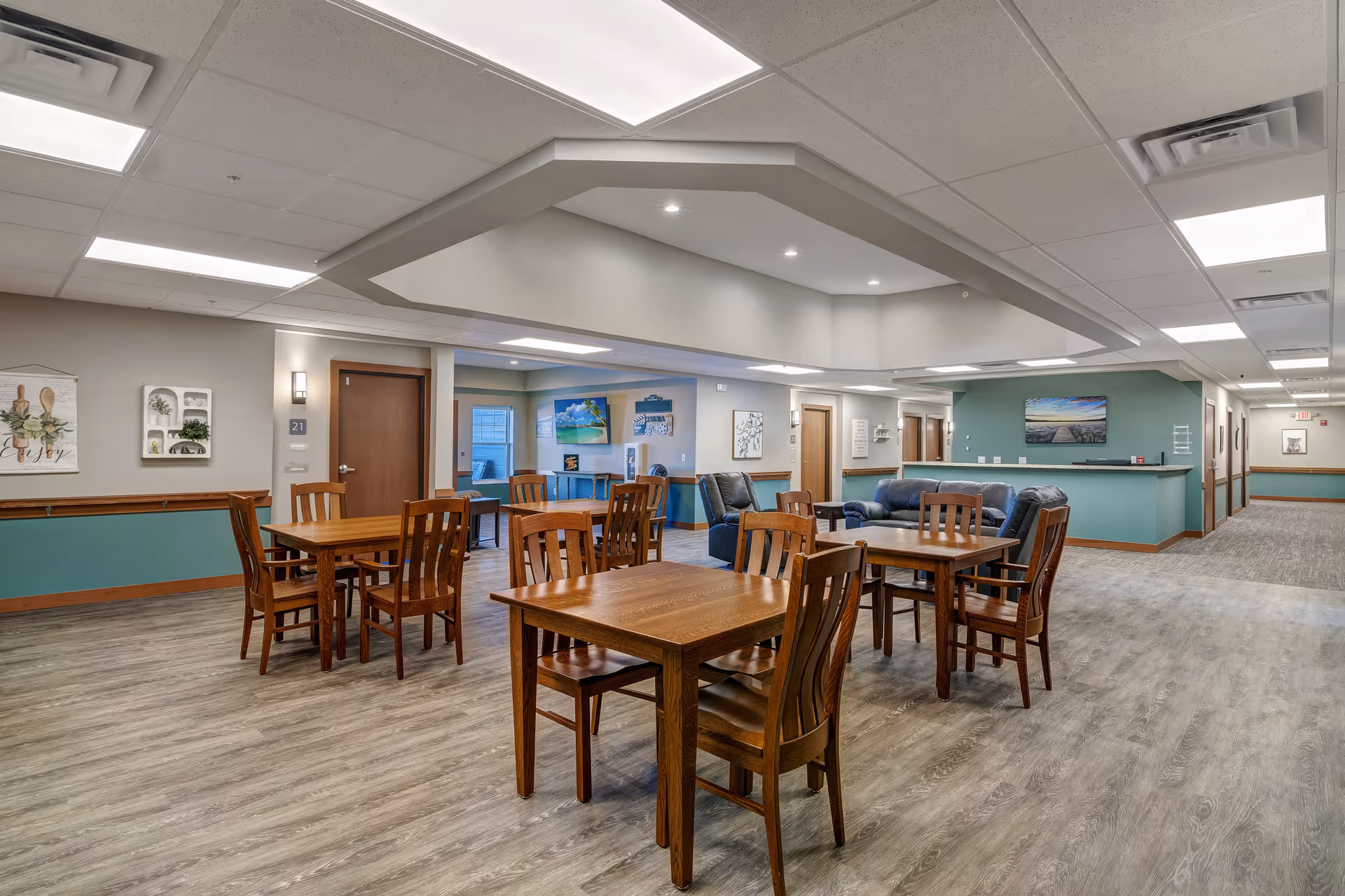 A spacious common area in a senior living facility featuring several wooden tables with chairs arranged around them. In the background, there is a seating area with blue leather sofas and armchairs. The walls are painted in neutral tones with teal accents, and there are various pieces of artwork and decor on the walls. The ceiling has recessed lighting and a unique architectural design element. Doors and hallways lead to other parts of the facility.