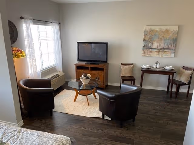 A cozy living area with two dark brown leather armchairs around a round glass coffee table on a light rug. A wooden TV stand with a flat-screen TV is against the wall. To the right, there is a small wooden table set with white dishes and two wooden chairs with cushions. A large window with sheer white curtains lets in natural light, and a piece of artwork hangs on the wall above the table.
