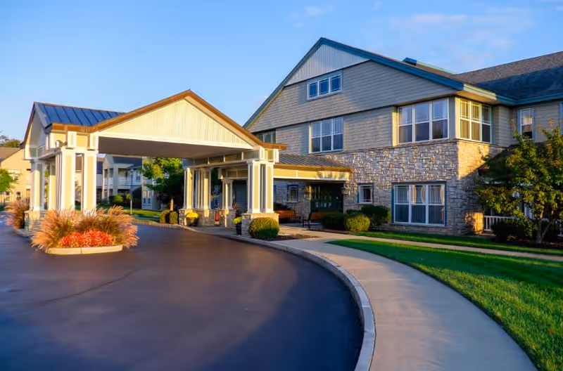 Exterior view of a senior living facility named Glen Eddy showing a covered entrance with white columns, a curved driveway, well-maintained landscaping with bushes and grass, and a multi-story building with stone and siding exterior under a clear blue sky.